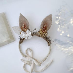 May include: A headband featuring two upright beige felt ears, decorated with dried flowers, small white fabric bows, and textured brown fibers. The band is wrapped in light fabric and includes long, thin fabric ties for securing the accessory.