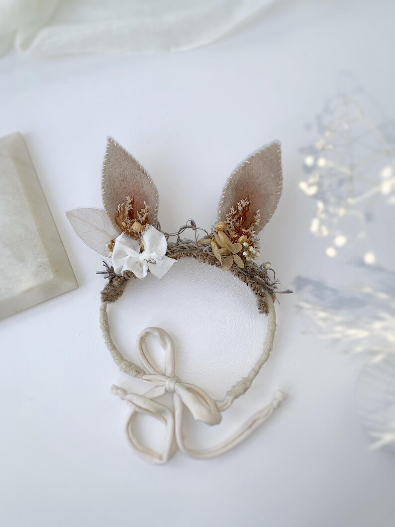 May include: A headband featuring two upright beige felt ears, decorated with dried flowers, small white fabric bows, and textured brown fibers. The band is wrapped in light fabric and includes long, thin fabric ties for securing the accessory.