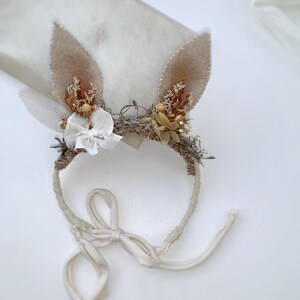 May include: A headband featuring two tan felt rabbit ears adorned with dried flowers, small white fabric blossoms, and a white leaf. The band is wrapped in textured material and includes long, cream-colored fabric ties for securing the accessory.