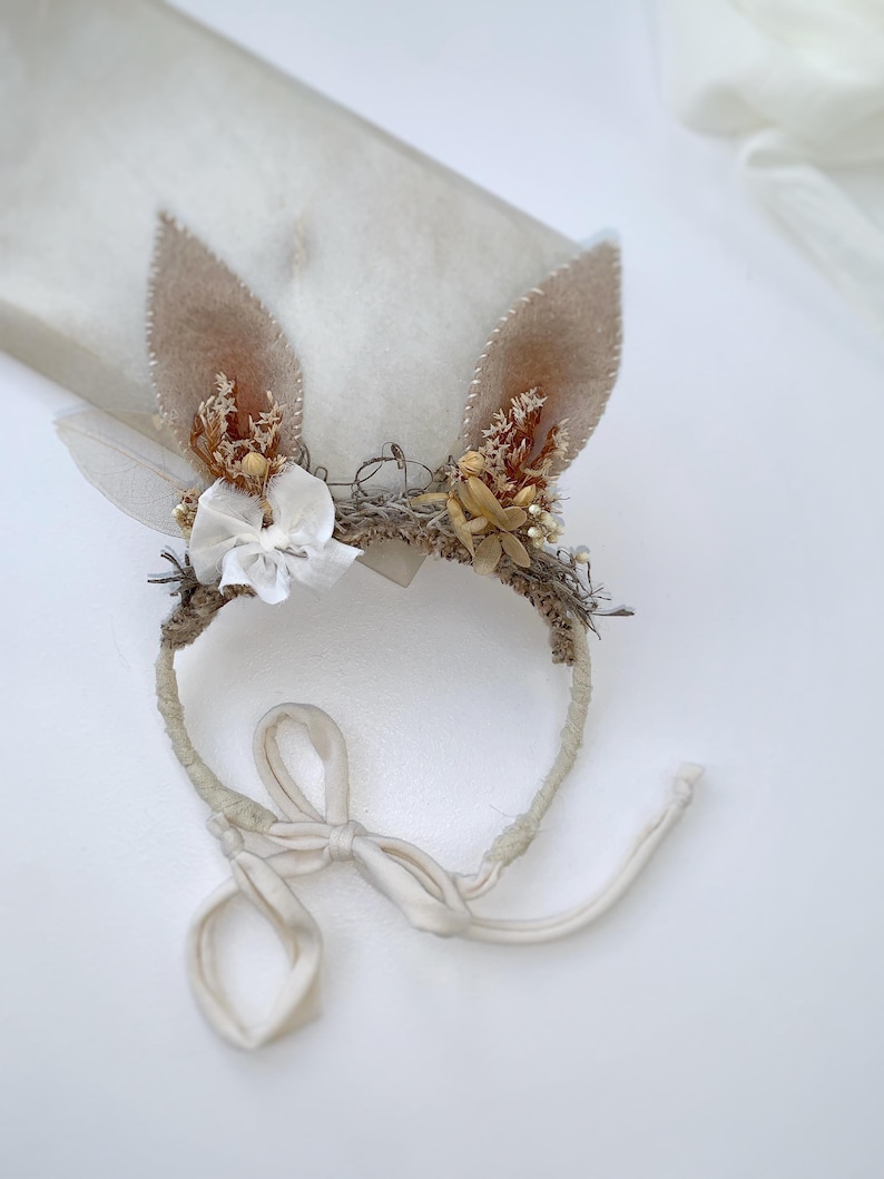 May include: A headband featuring two tan felt rabbit ears adorned with dried flowers, small white fabric blossoms, and a white leaf. The band is wrapped in textured material and includes long, cream-colored fabric ties for securing the accessory.