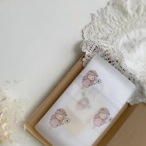 May include: A rectangular cardboard box containing white tissue paper printed with a repeating illustration of a girl with long brown hair wearing a pink dress and a flower crown, holding a dandelion. The text reads SORI'S LITTLE CUTIES.
