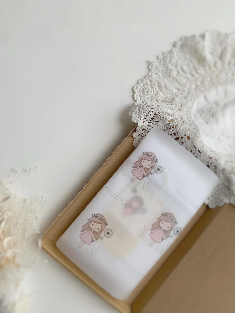 May include: A rectangular cardboard box containing white tissue paper printed with a repeating illustration of a girl with long brown hair wearing a pink dress and a flower crown, holding a dandelion. The text reads SORI'S LITTLE CUTIES.