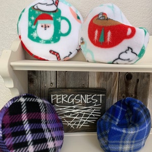 May include: Four plush, round, and beanbag-style pillows with various winter-themed patterns. The pillows are arranged on a wooden shelf with a sign that reads "Fergsnest". The pillows feature a red and white mug with a snowman, a red and white mug with a Christmas tree, a purple and black plaid pattern, and a blue and white plaid pattern.