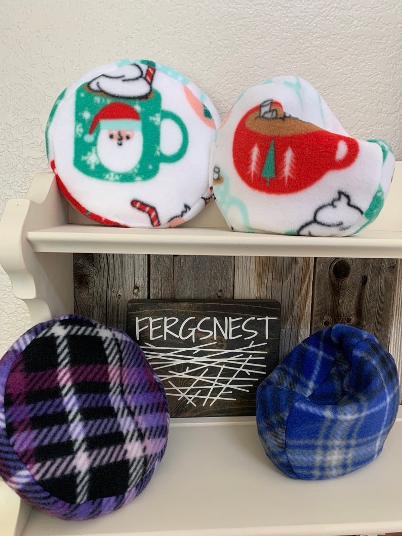 May include: Four plush, round, and beanbag-style pillows with various winter-themed patterns. The pillows are arranged on a wooden shelf with a sign that reads "Fergsnest". The pillows feature a red and white mug with a snowman, a red and white mug with a Christmas tree, a purple and black plaid pattern, and a blue and white plaid pattern.