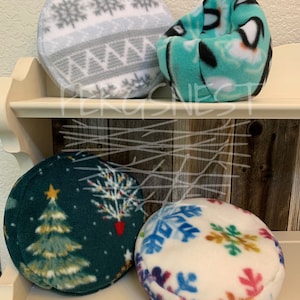 May include: Four round fleece pet beds in various winter holiday patterns. The beds are on a wooden shelf. The patterns include snowflakes, a Christmas tree, and a gray and white geometric pattern.