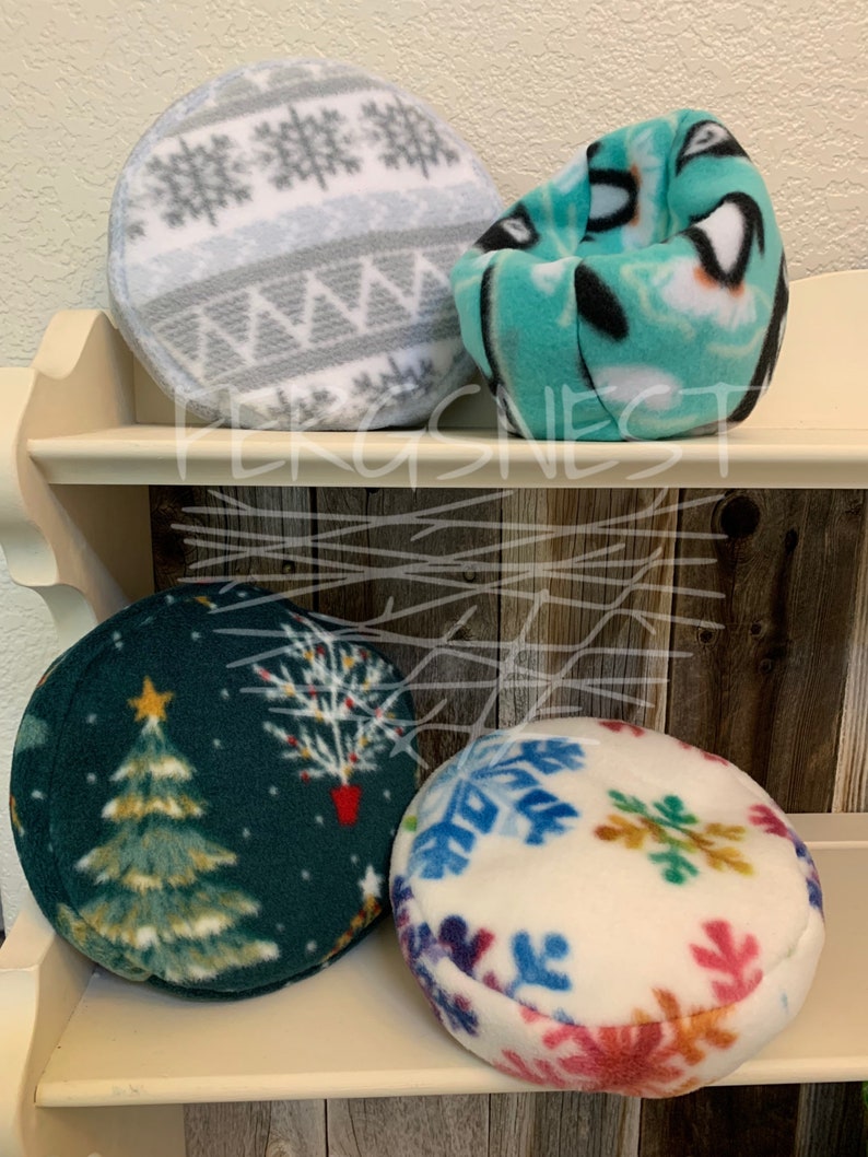 May include: Four round fleece pet beds in various winter holiday patterns. The beds are on a wooden shelf. The patterns include snowflakes, a Christmas tree, and a gray and white geometric pattern.