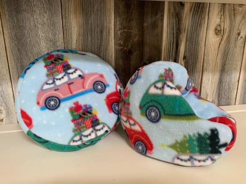 May include: Two blue fleece Christmas-themed car pillows. One is round and the other is shaped like a foot. Both feature a light blue background with red, green, and white cars with Christmas trees and presents on top.