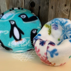 May include: Two fleece pet beds, one with a turquoise background and a penguin pattern, the other with a white background and a blue and purple snowflake pattern. The text "Fergsnest" is visible on the penguin bed.