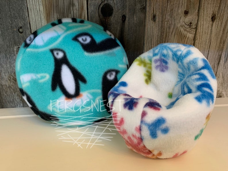 May include: Two fleece pet beds, one with a turquoise background and a penguin pattern, the other with a white background and a blue and purple snowflake pattern. The text "Fergsnest" is visible on the penguin bed.