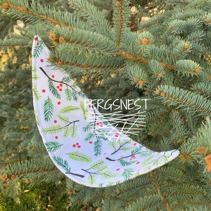 May include: A white fabric crescent moon with a green and red holly berry pattern hanging from a tree branch. The fabric has a white background with green holly leaves and red berries.