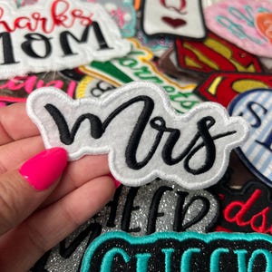 May include: A collection of embroidered patches with various designs and text. The central patch reads "Mrs" in black script on a white background with a white border. Other patches include the words "Sharks Mom", "Princess", and "Cheer".