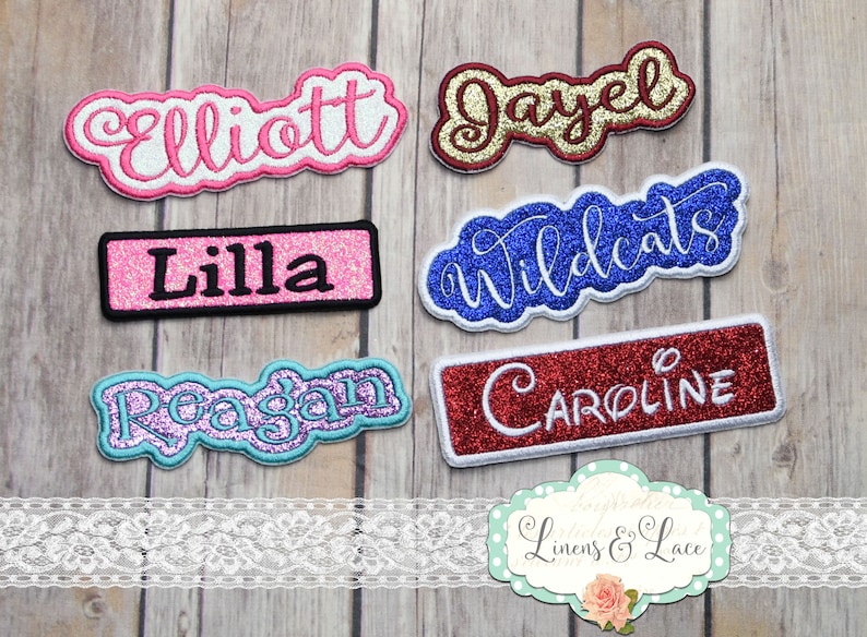 May include: Embroidered name patches in a variety of colours and styles. Names include "Elliott," "Jayel," "Lilla," "Wildcats," "Reagan," and "Caroline." Each patch has a glittery or textured design with contrasting borders.