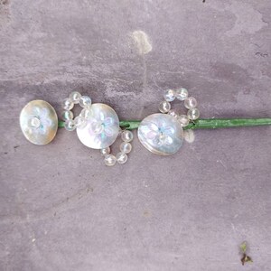 May include: Three iridescent white shell buttons with a floral design, strung on a clear beaded strand with a green stem.
