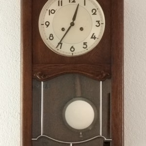 May include: A rectangular, wooden wall clock with a cream-colored clock face and black numerals. The clock has a pendulum visible through a glass panel. The wooden frame is a dark brown color.