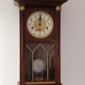 Puede incluir: Un reloj de pared vintage de madera con una esfera blanca y marcadores de hora romanos negros. El reloj tiene un frente de vidrio y un péndulo en el interior.
