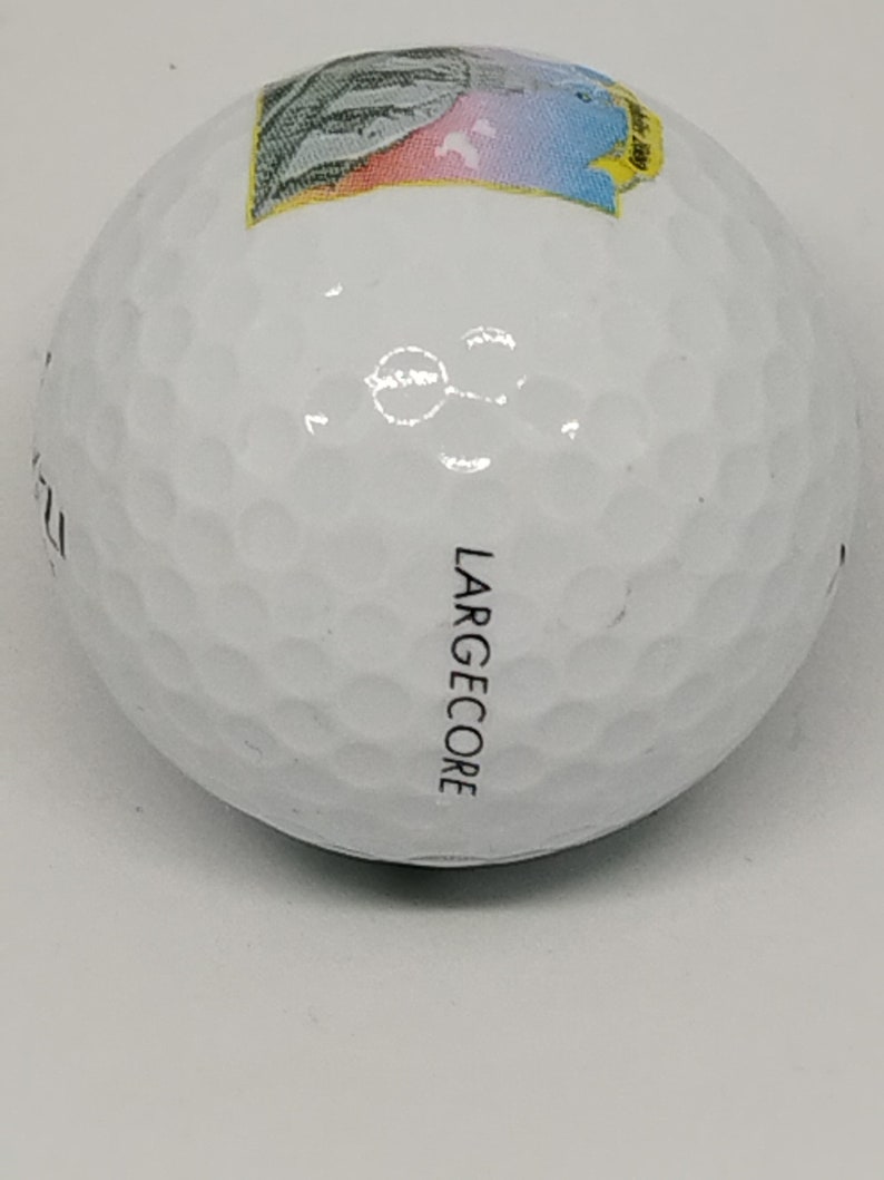 May include: White golf ball with a colorful graphic on one side and the word "LARGE CORE" printed on the other side.