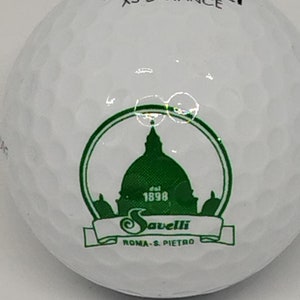 May include: White golf ball with a green logo featuring a building with a dome and the text "Savelli Roma - S. Pietro" and "Est. 1898". The text "Maxfli XS Distance" is printed on the top of the ball.