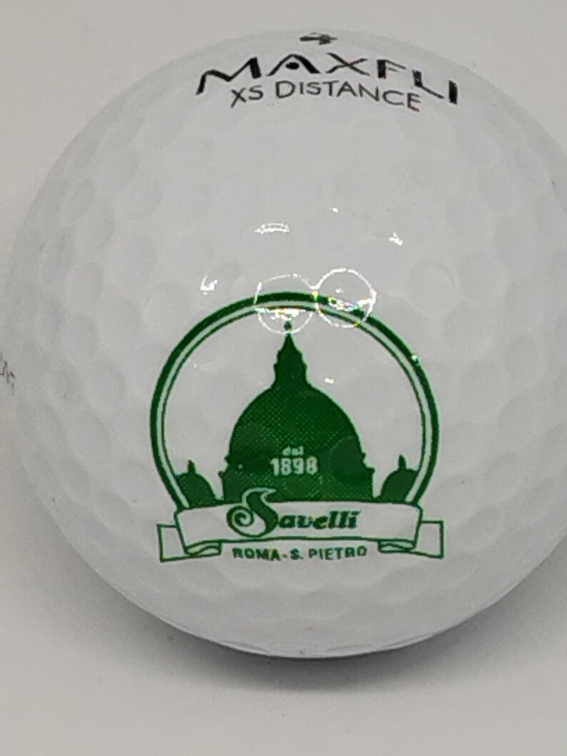 May include: White golf ball with a green logo featuring a building with a dome and the text "Savelli Roma - S. Pietro" and "Est. 1898". The text "Maxfli XS Distance" is printed on the top of the ball.