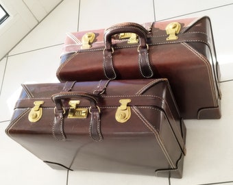 Op de afbeelding: Twee vintage bruin lederen koffers, op elkaar gestapeld. De koffers hebben messing hardware, waaronder sloten en handvatten. Witte stiksels accentueren de randen van de bagage en de handvatten zijn donkerbruin.