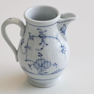 Puede incluir: Una jarra de cerámica blanca con un patrón floral azul. La jarra tiene un cuerpo redondeado y un pequeño pico.