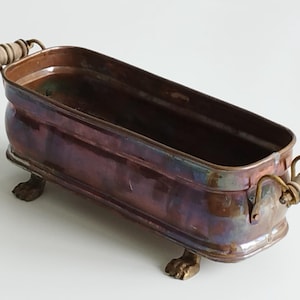May include: A copper planter with wooden handles and brass feet. The planter has an iridescent finish and is oval shaped.