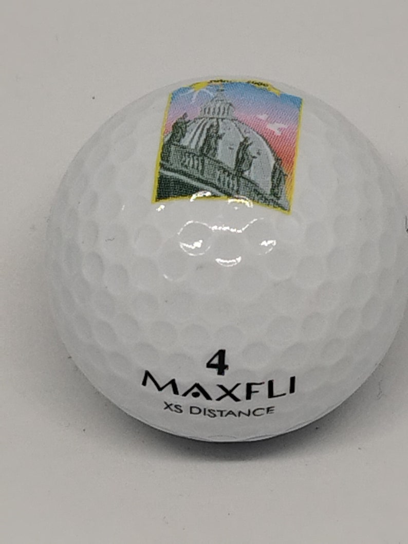May include: White golf ball with a black and white image of a building with a dome on top. The ball has the number 4 and the text "MAXFLI XS DISTANCE" printed on it.