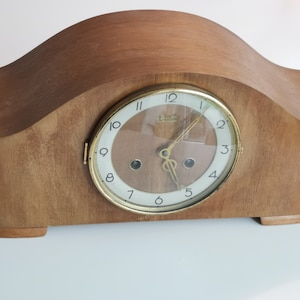 Puede incluir: Un reloj de chimenea de madera con una esfera dorada y marcadores de hora romanos negros. El reloj tiene un fondo blanco con un centro marrón y una cubierta de vidrio. La esfera del reloj dice "Elma"