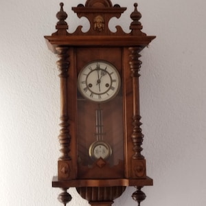 Könnte beinhalten: Eine Wanduhr aus Holz mit Pendel. Das Zifferblatt ist hell mit schwarzen römischen Ziffern und Zeigern. Der Holzrahmen ist aufwendig mit dekorativen Elementen und einem kleinen Gesicht oben geschnitzt. Die Uhr ist an einer weißen Wand befestigt.
