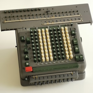 Op de afbeelding: Een vintage grijze mechanische rekenmachine met een groen toetsenbord en een schuifwagen. De rekenmachine heeft een reeks knoppen met de cijfers 1 tot en met 9 en een paar functietoetsen, waaronder "STOP", "Div", "+" en "-".