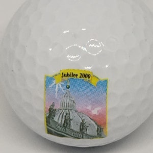 May include: White golf ball with a black Maxfli logo and the number 4 printed on it. The ball also has a colorful sticker with the text "Jubilee 2000" and a picture of a building.