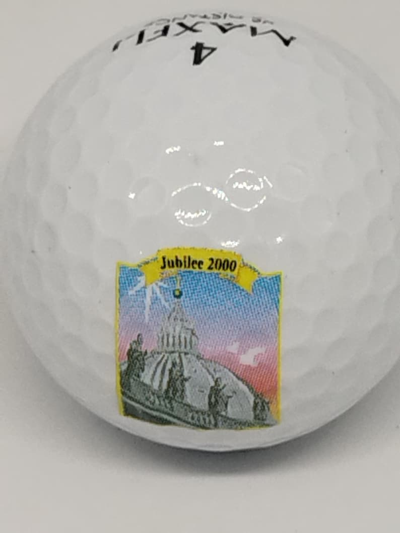 May include: White golf ball with a black Maxfli logo and the number 4 printed on it. The ball also has a colorful sticker with the text "Jubilee 2000" and a picture of a building.
