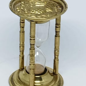 May include: A brass hourglass with an ornate top and a clear glass sand timer. The hourglass is on a white background.