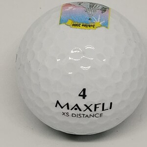 May include: White golf ball with black text that reads "4 MAXFLI XS DISTANCE" and a small sticker on the top that reads "Jubilee 2000".