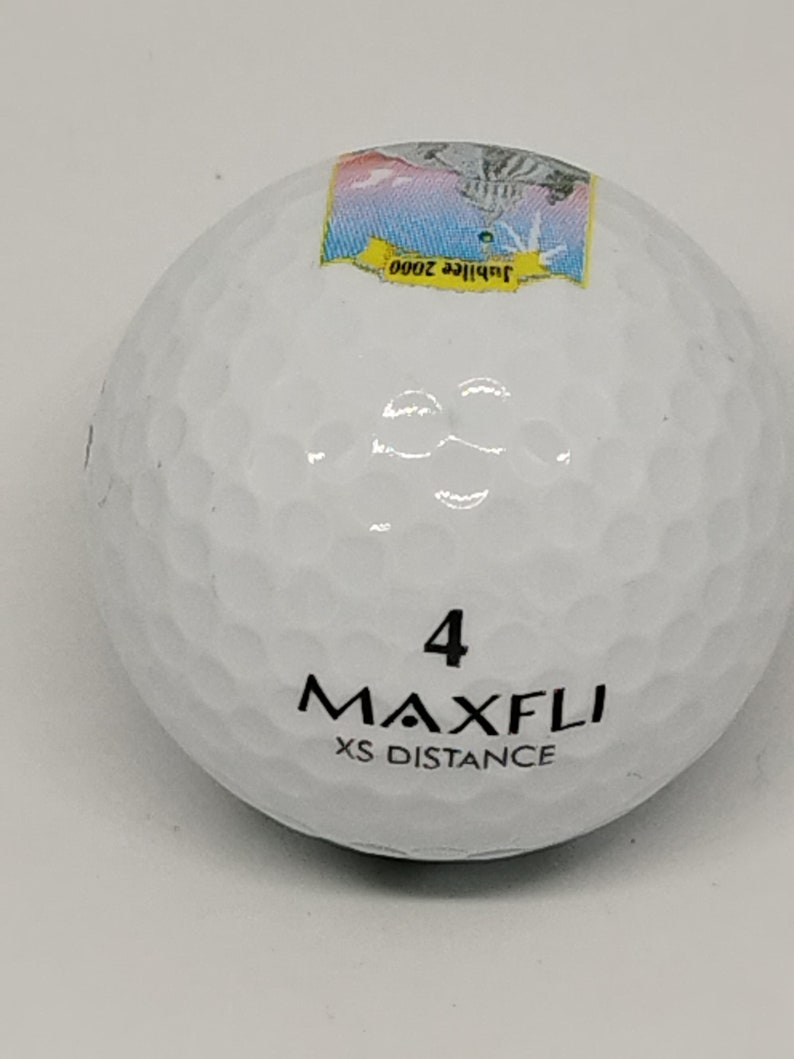 May include: White golf ball with black text that reads "4 MAXFLI XS DISTANCE" and a small sticker on the top that reads "Jubilee 2000".