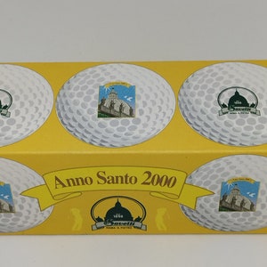May include: A yellow box with four white golf balls. Each golf ball has a different image of a building with a dome. The box says "Anno Santo 2000" and "Savelli Roma & S. Pietro" in green and white.