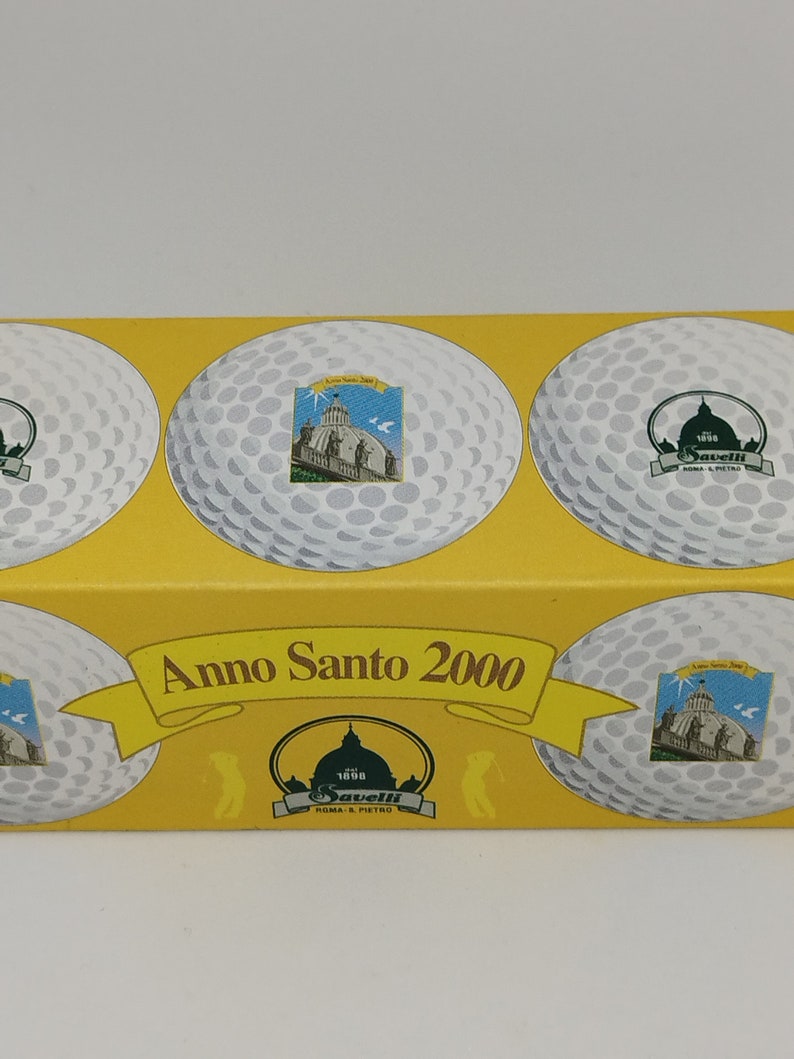 May include: A yellow box with four white golf balls. Each golf ball has a different image of a building with a dome. The box says "Anno Santo 2000" and "Savelli Roma & S. Pietro" in green and white.