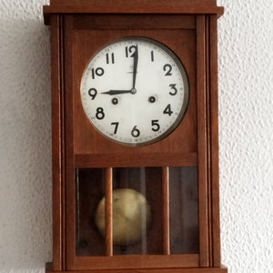 Puede incluir: Un reloj antiguo de madera con una esfera blanca y agujas negras. El reloj tiene un péndulo y está colgado en una pared blanca.