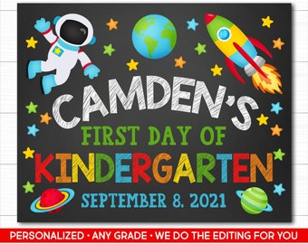 Puede incluir: Un letrero de pizarra negra con gráficos del espacio que incluyen un astronauta, un cohete, la Tierra y planetas. El texto dice CAMDEN'S FIRST DAY OF KINDERGARTEN SEPTEMBER 8, 2021 en letras coloridas de estilo dibujado a mano con estrellas amarillas.