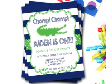 May include: A stack of navy blue and white striped invitations. Each invitation features a green alligator graphic and the text "Chomp! Chomp! AIDEN IS ONE!" with event details. Colorful confetti and straws are in the background.