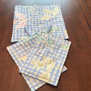 May include: Set of four blue and white quilted pot holders with a floral pattern. The pot holders are made of fabric and have a checkered pattern.
