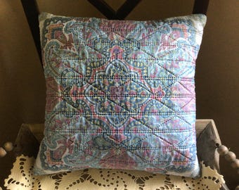 May include: Square decorative pillow with a quilted paisley pattern in shades of blue, pink, and green. The pillow is placed on a wooden chair and has a white border.