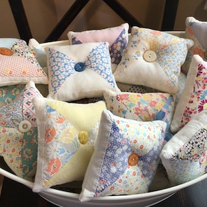 May include: A collection of small, square, handmade pillows with floral and patterned fabric designs. Each pillow features a button in the center and is filled with a soft material. The pillows are in a white bowl.