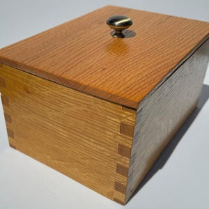 May include: A wooden box with a hinged lid and a metal knob. The box is made of light brown wood with a grain pattern. The lid is closed.