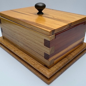 Peut inclure: Une boîte en bois avec un couvercle à charnières et un bouton foncé. La boîte est faite de différents types de bois, dont l'érable, le cerisier et le noyer. La boîte est posée sur une base en bois.