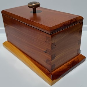 Peut inclure: Une boîte en bois avec un couvercle à charnières et un bouton en laiton. La boîte est en bois de cèdre et a une couleur brun rougeâtre naturelle. La boîte est posée sur une base en bois.