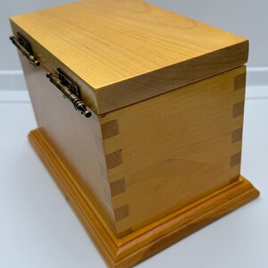 May include: A wooden box with a hinged lid and brass hinges. The box is made of light wood and has a natural finish. The box is sitting on a white surface.