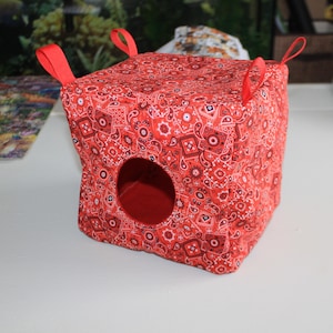 May include: A red bandana-print cube-shaped pet bed with a circular opening and red fabric lining. The bed has four red fabric loops at the corners. The fabric features a paisley pattern in red, white, and black.