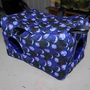 May include: A blue and black fabric pet hammock with a moon and star pattern. The hammock has two openings and is designed for small animals.