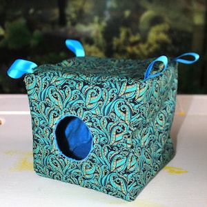 Peut inclure: Un cube en tissu avec un motif de tentacules de pieuvre turquoise et noir. Le cube présente une ouverture circulaire sur la face avant, révélant une doublure intérieure bleu uni. Quatre boucles en ruban bleu vif sont fixées aux coins supérieurs.