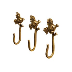 Puede incluir: Tres ganchos de pared de metal color latón, cada uno con una figura de rana esculpida en la parte superior. La rana se muestra en posición de escalada con las extremidades extendidas, mientras que la parte inferior forma un gancho en forma de J.
